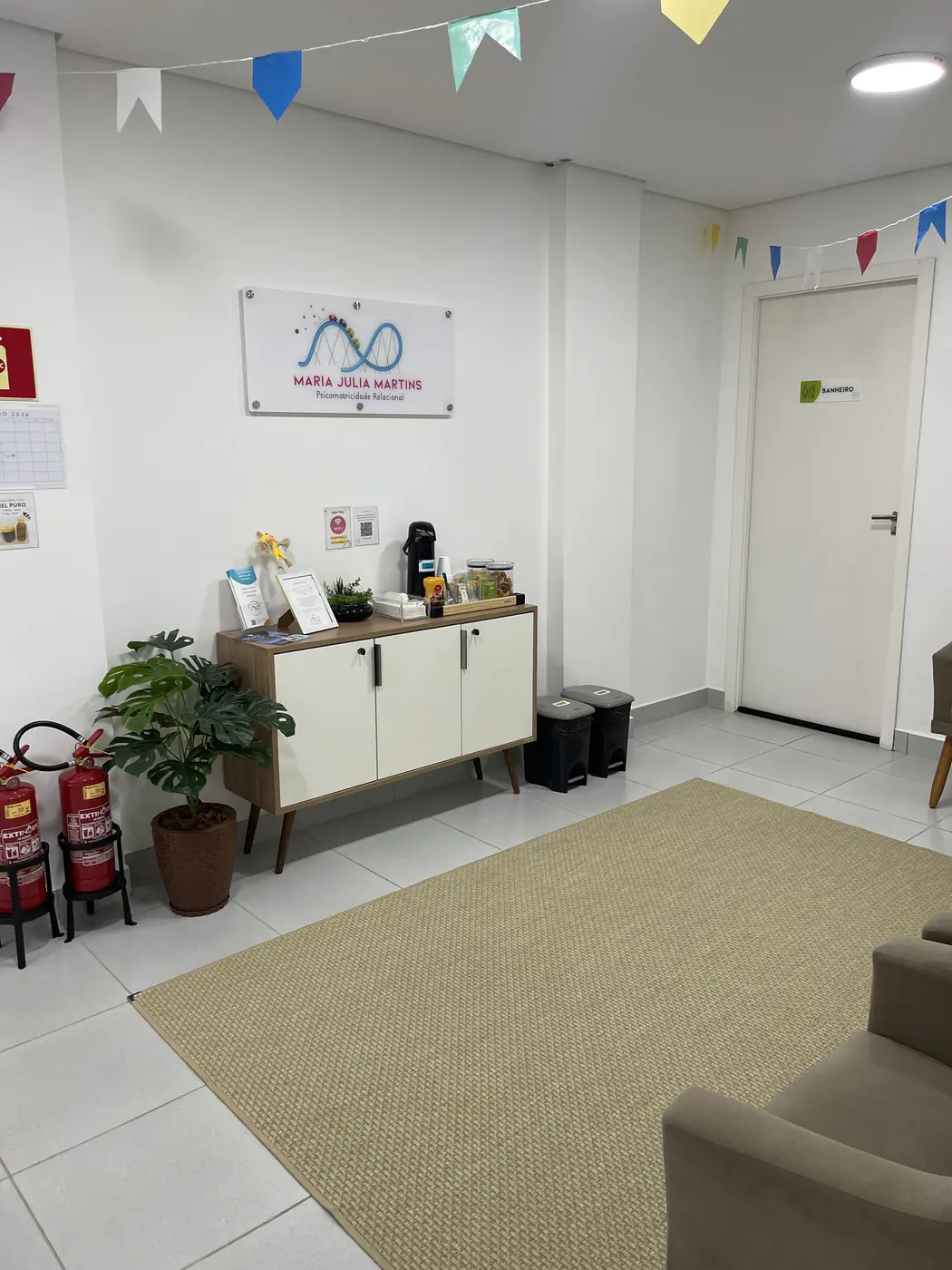 Recepção com o logotipo Maria Julia Martins na parede, aparador, plantas e bandeirinhas coloridas