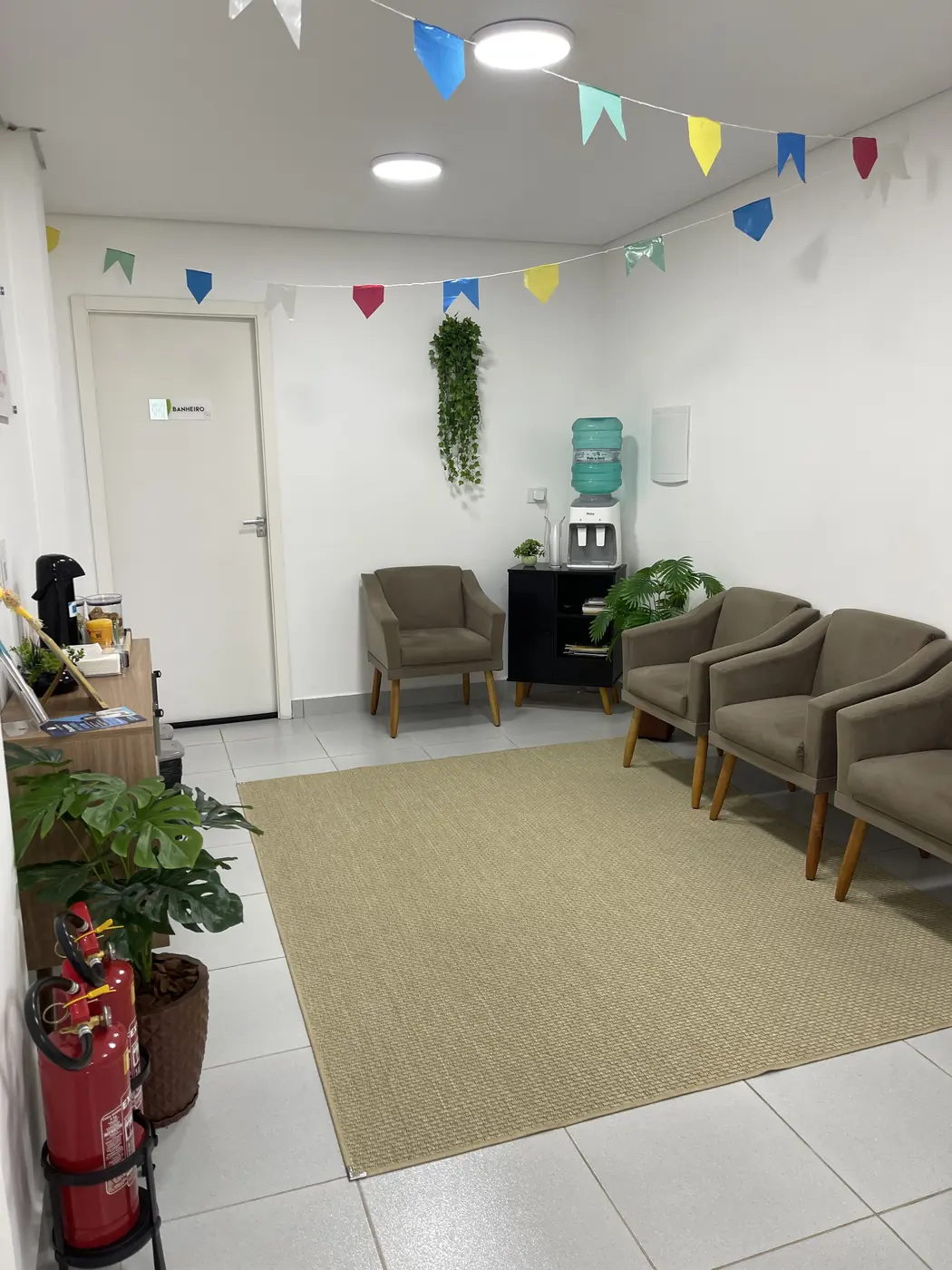 Sala de espera da clínica com poltronas, tapete, bandeirinhas coloridas e bebedouro